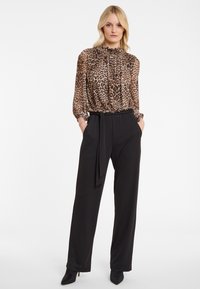 Woman wearing a high-neck leopard print blouse with long sleeves tucked into high-waisted black wide-leg trousers with a waist tie.