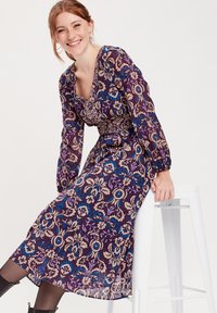 Vestido largo morado con patrones florales en beige, azul y púrpura claro. Presenta un escote profundo y cintura ajustada con mangas abullonadas.