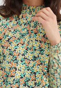 Blusa floreale con sfondo nero, caratterizzata da fiori gialli e arancioni, scollatura arricciata e dettagli con polsini elasticizzati.