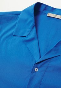 Chemise boutonnée bleu vif avec un col classique et un bouton blanc visible, posée à plat sur un fond blanc.