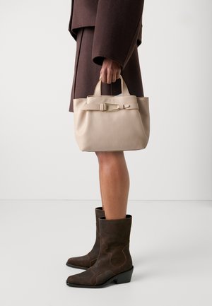 Borsa in pelle beige con manici corti, caratterizzata da una fibbia in tono oro. Il modello indossa un cappotto oversize marrone e stivaletti alla caviglia marrone scuro.