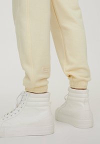 Kremowe dresy z elastycznymi mankietami i małą metką, w połączeniu z białymi sneakersami typu high-top z teksturowanymi akcentami i płaską podeszwą.