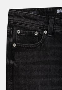 Närbild av svarta denimjeans som visar frontfickan med silvernitar och bältesöggla, med etiketten "Relaxed Chris" inuti midjan.