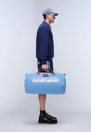 Homme avec une casquette bleue, des lunettes de soleil et une tenue, portant un sac de voyage Napapijri bleu clair à la main, debout devant un fond clair uni.