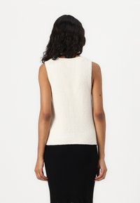 Beżowa, bezrękawnikowa bluzka dzianinowa o teksturowanej tkaninie, okrągłym dekolcie i dopasowanym kroju. Brak widocznych elementów metalowych lub wzorów.