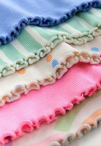 Ribbet stof i blå, grønstribet, creme med multifarvede prikker og pink, alle med flæsekant og blød tekstur. Hvert lag er stablet ovenpå hinanden.