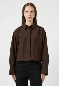 Camicia corta marrone in cotone con chiusura a bottoni, colletto a punta e due tasche frontali. Maniche lunghe con vestibilità leggermente ampia.