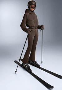 Hnedý lyžiarsky oblek s priliehavým strihom, ktorý obsahuje kapucňu, dve predné vrecká a opasok. Čierne lyže, lyžiarske topánky a paličky dopĺňajú tento outfit.