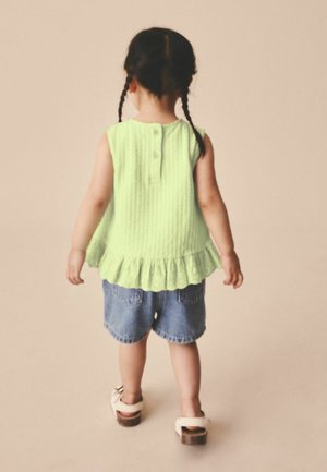Toddler with braided hair wearing a sleeveless green top, denim shorts, and white sandals standing facing away against beige background.