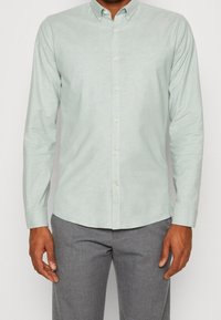 Jasnozielona koszula zapinana na guziki, wykonana z miękkiego, teksturowanego materiału, z kołnierzykiem i długimi rękawami, w połączeniu z szarymi spodniami.