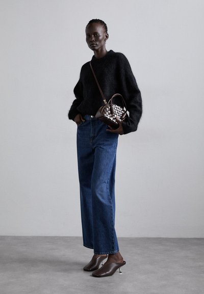 Zwarte fuzzy trui, high-waisted blauwe denim flared pants, bruin geweven handtas en bruine muiltjes met een metalen hak. Effen achtergrond.