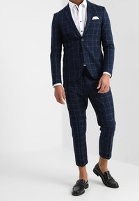 Navyblauer karierten Anzug mit slim-fit Design. Enthält ein weißes Hemd, ein Einstecktuch und schwarze Slipper. Maßgeschneiderte Hosen mit cropped Saum.