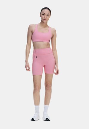 Junge Frau trägt einen pinken Sport-BH und passende hoch taillierte Biker-Shorts, weiße Socken und weiße Sportschuhe und steht vor einem einfarbigen Hintergrund.