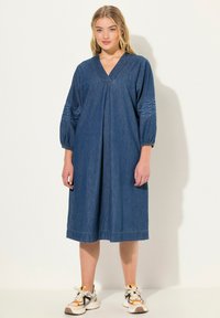 Denimmekko, jossa on V-pääntie, kolmanneksen hihat ja broderoidut yksityiskohdat. A-linjan siluetti polvipituudella ja hieman rento istuvuus.