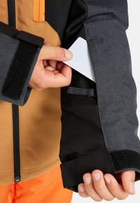 Wasserabweisende Jacke in Schwarz und Orange, ausgestattet mit einer Kartenasche am Ärmel und verstellbaren Klettverschluss-Manschetten.