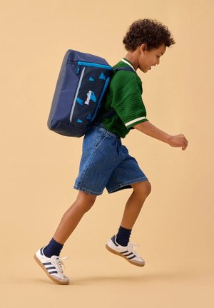 Marineblaue Rucksack mit türkisfarbenen Akzenten und geometrischen Mustern. Getragen von einem Kind in einem grünen Pullover und Jeansshorts, das verspielt springt.