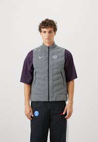 Gray quilted vest with high collar, front zipper, and side pockets, worn over a purple short-sleeve shirt, paired with black pants.