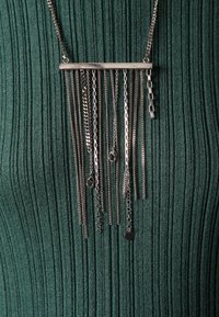 Collier en argent avec différents styles de chaînes suspendus verticalement à une barre horizontale sur un fond de tissu côtelé vert foncé.