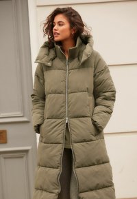 Manteau d'hiver long vert olive avec fermeture éclair, capuche surdimensionnée et design matelassé. Il dispose de poches latérales et d'un col côtelé.