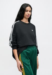 Čierna mikina s bielymi pruhmi na rukávoch a malým logom; kombinovaná so zelenými nohavicami s žltými bočnými pruhmi; voľný strih.