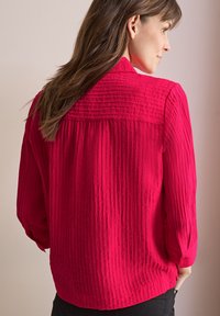 Camicia stropicciata di un vivace colore rosa con colletto, maniche lunghe e polsini abbottonati, caratterizzata da linee verticali testurizzate e orlo curvo.