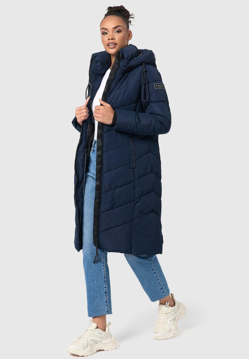 Doudoune bleu marine avec capuche, présentant une fermeture éclair sur le devant et un motif matelassé en diagonale. Associée à un jean bleu clair et des baskets blanches.