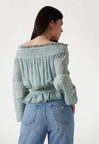 Mint green off-shoulder blouse featuring smocked bodice, lace sleeves, and ruffled hem, paired with light blue denim jeans.
