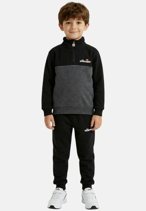 Un garçon se tient face à l'avant, portant un pull à manches longues noir et gris, un pantalon noir et des baskets blanches, tous ornés de logos ellesse.
