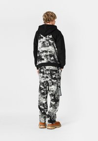 Sudadera con patrón en blanco y negro, con la parte delantera negra y un diseño abstracto en la parte trasera, combinada con pantalones holgados a juego y calzado marrón.