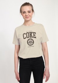Beige Baumwoll-T-Shirt mit schwarzem "COKE"-Druck und Atlanta-Branding, kurze Ärmel, lockere Passform, kombiniert mit schwarzen Jeans, einfaches Design.