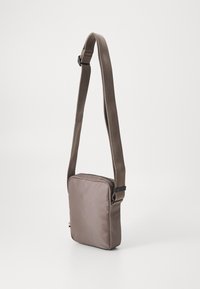 Bolso bandolera de tela marrón con forma rectangular, correa ajustable y cierre de cremallera. Presenta una superficie texturizada y un diseño minimalista.