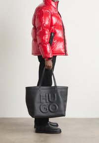 Rote Steppjacke mit glänzendem Finish, schwarze Akzente, kombiniert mit einer großen schwarzen Leder-Handtasche mit eingeprägtem "HUGO"-Logo. Schwarze Hose und Schuhe.