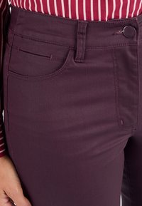 Jeans de mezclilla burdeos con una textura suave, que presentan un bolsillo frontal, trabillas para el cinturón y un cierre de botón único en la cintura.