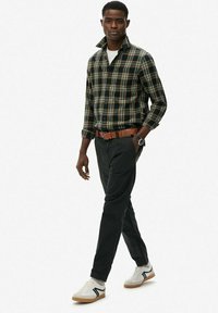 Mand, der går, iført grøn ternet skjorte med opretstående krave, sorte bukser, brun bælte, hvide sneakers og armbåndsur på venstre håndled.