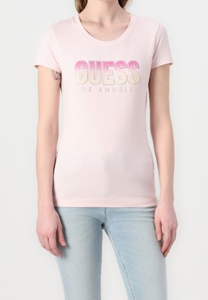 Femme portant un t-shirt Guess Los Angeles rose clair associé à un jean bleu clair, debout devant un fond uni.