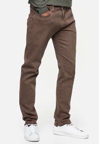 Pantalones de algodón marrón con corte slim, que cuentan con bolsillos frontales y una cremallera en la bragueta, combinados con zapatillas blancas. Textura suave y diseño limpio.