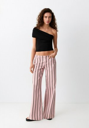 Žena s kovrčavom kosom koja nosi crni crop top s jednim ramenom i ružičaste široke hlače s okomitim crnim prugama, stoji uz bijeli zid.