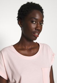 Lichtroze T-shirt met korte mouwen, gemaakt van zachte stof, met een ronde halslijn en een enkele borstzak. Platte naaddetails op de mouwen.