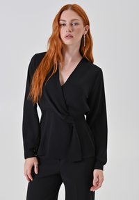 Blusa nera avvolgente con maniche lunghe, scollatura a V e dettaglio con cintura. Tessuto liscio, silhouette aderente e sottili accenti con bottoni sulle maniche.