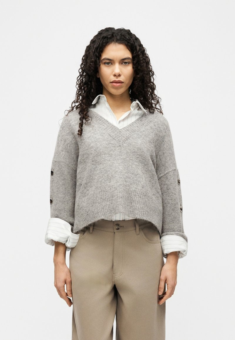 Γυναίκα με σγουρά μαλλιά φοράει γκρι πουλόβερ με V λαιμόκοψη πάνω από λευκό πουκάμισο με γιακά και μπεζ παντελόνι, στέκεται μπροστά σε απλό φωτεινό φόντο.