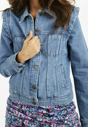 Femme portant une veste en jean bleue avec des boutons métalliques et une jupe à motif floral, tenant le col de la veste d'une main.