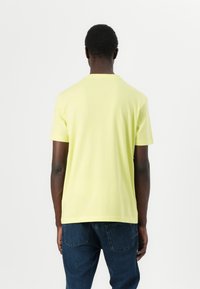 Korte ermer, lys gul t-skjorte laget av bomull med rund hals, vist fra baksiden, sammen med mørkeblå jeans.
