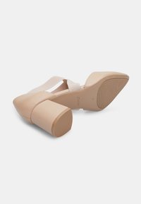 Beige high-heeled shoe with a pointed toe and a wide block heel. Features elastic straps across the top, smooth leather texture, and rubber sole.