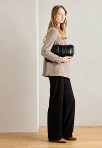 Sac à main noir plissé tenu sous le bras, avec une texture lisse. Le mannequin porte un blazer à carreaux et un pantalon noir à jambes larges. Fond en parquet.