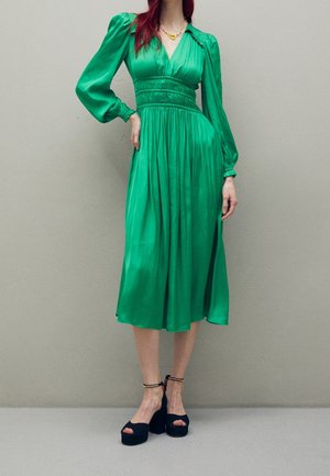 Vrouw met een emerald groene midi-jurk met lange mouwen en een getailleerde taille, gecombineerd met zwarte plateauhakken met enkelbandjes.