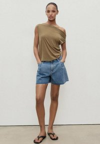 Brun, enaxlad topp; blå denimshorts med fransad nederkant; svarta sandaler med nitar; neutral bakgrund; modellen står i en avslappnad pose.