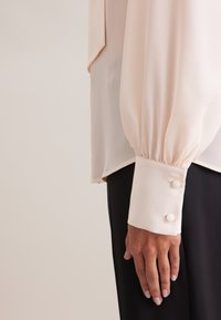 Blouse with puffed long sleeves in pale pink fabric. Features buttoned cuffs and a smooth, lightweight texture. Positioned with a dark backdrop.