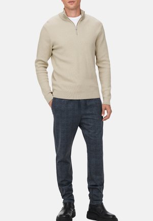 Beige gebreide trui met een kwart rits en lange mouwen, gecombineerd met donkergepatenteerde broeken en zwarte enkellaarzen. Textuurdetails in de stof zijn zichtbaar.