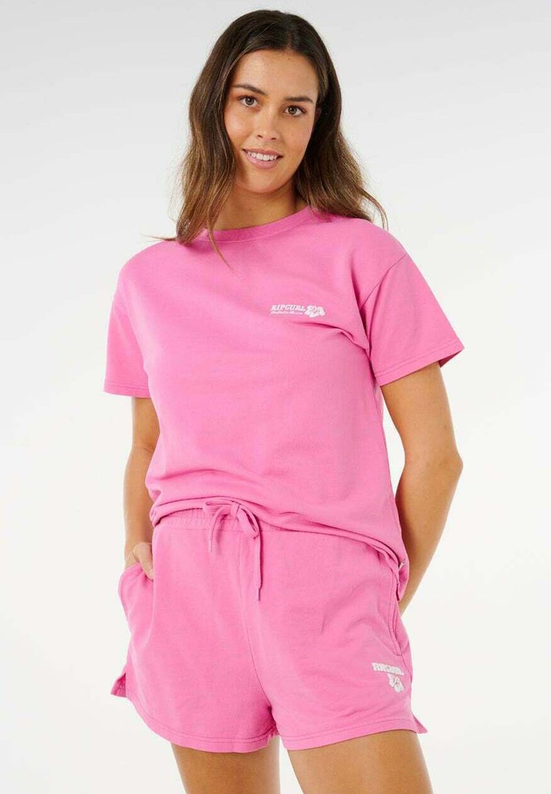 Frau, die lächelt und ein hellpinkes T-Shirt mit kurzen Ärmeln sowie passende Shorts mit Kordelzug trägt, jeweils mit kleinen weißen Logos auf der Brust und am Bein.