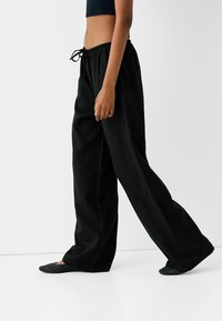 Pantalons larges noirs avec une taille élastique et un cordon de serrage. Le tissu semble léger avec une texture lisse, associé à des chaussures plates en toile.